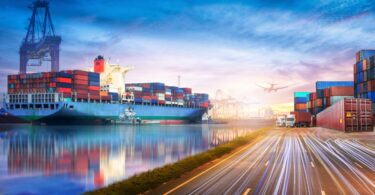 A fully loaded cargo ship in the ocean next to a busy road and a freight plane flying overhead.