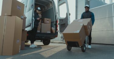 Workers in safety gear loading cardboard boxes on a dolly from a commercial van, with a female worker organizing inventory.