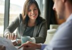 A woman in a blazer smiling as she reaches over a table to point to something on a paper her male client is holding.