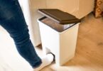 A person wearing blue jeans and white socks opens a white trashcan using their foot.