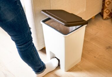 A person wearing blue jeans and white socks opens a white trashcan using their foot.