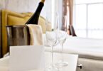 A bucket of ice with a bottle of champagne in it sitting beside two glasses on a hotel room’s bedside table.