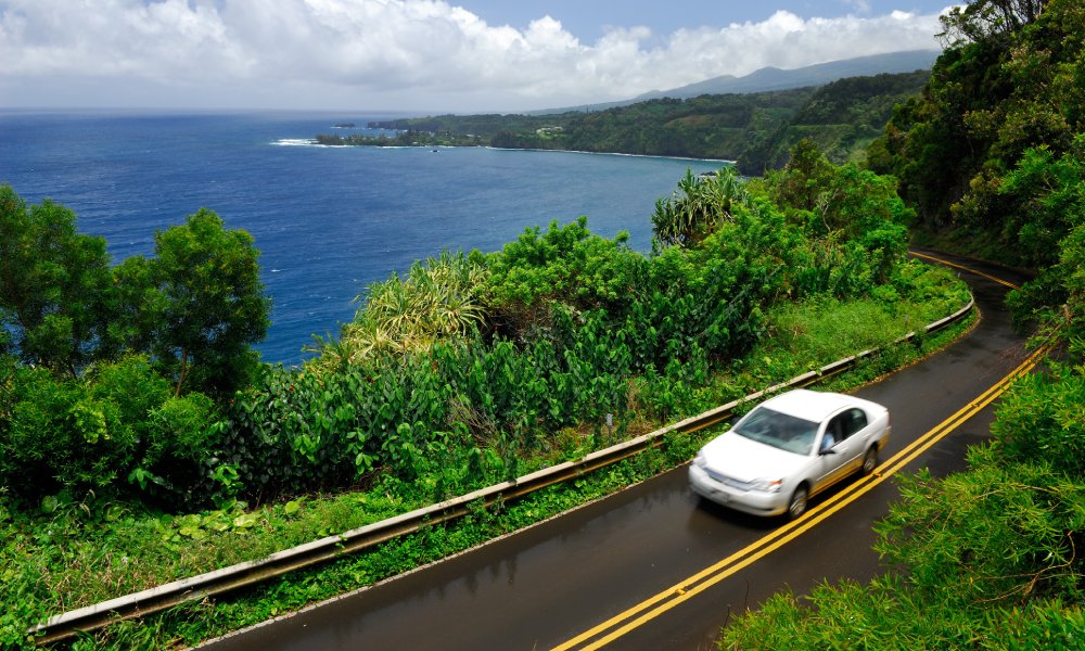 A white car drives on a two-lane stretch of the Hana Highway in Maui, Hawaii, with the ocean in the background.