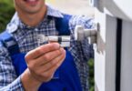 A locksmith in a blue and white plaid shirt holding a key and a cylinder lock outside a white door, exemplifying what to check before calling a 24/7 locksmith.