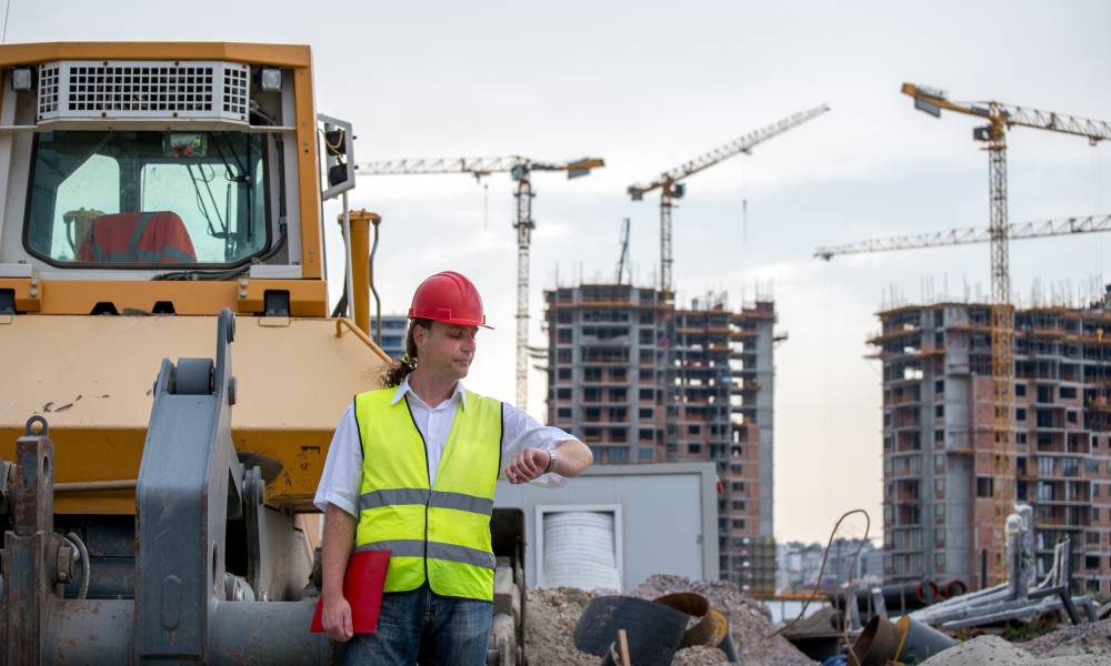 A construction manager stands on a jobsite and looks at his watch. Cranes and heavy equipment are in the background.