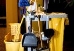 A collection of essential cleaning supplies for businesses displayed in a dimly lit room, including mops, buckets, brooms, and various cleaning agents on a janitorial cart.