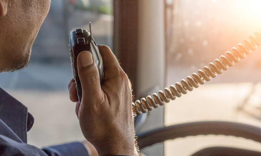 A close-up of a trucker holding a CB radio to his face to talk. Sunlight is streaming through his truck's windshield.
