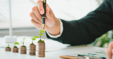 A row of increasingly taller coin stacks, each with a tiny plant on top. A hand points to the largest stack with a pen.