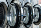 Four steel front-loaded laundry machines in a laundry facility with their round doors swung open.
