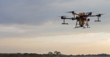 A black, multi-rotor drone flying above the pavement. It's carrying a beige tank in its center.