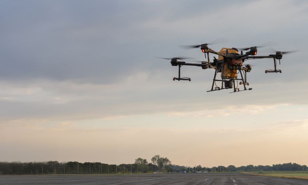 A black, multi-rotor drone flying above the pavement. It's carrying a beige tank in its center.