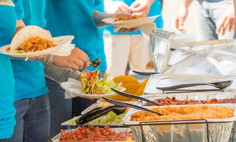 A close-up of people preparing tacos at a taco bar. The bar includes rice, beans, and various taco toppings.