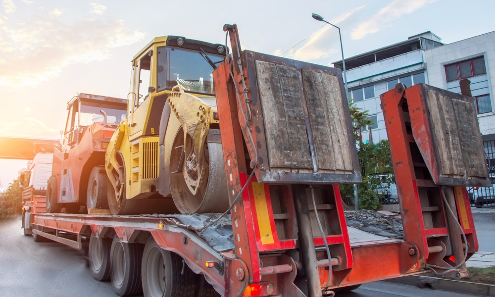 A semi truck transports two asphalt compactors on a flatbed trailer. The truck drives down the highway at sunset.