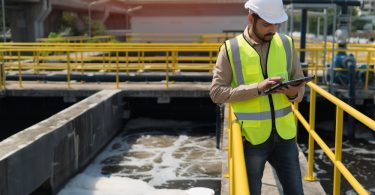 A man in a yellow safety vest and white hard hat looks at a tablet and checks on waste water treatment at a waste plant.