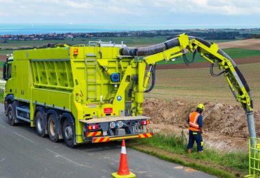 A worker supervising a neon yellow-green soil vacuum excavation truck with a hose going into the ground next to a road.