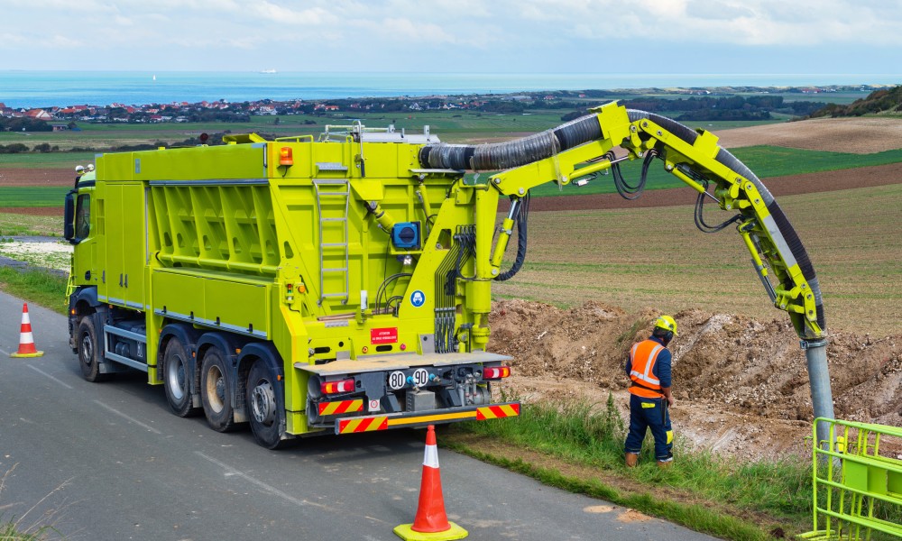 A worker supervising a neon yellow-green soil vacuum excavation truck with a hose going into the ground next to a road.