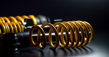 Three yellow springs and black shock absorber coil-overs rest on a black surface illuminated by studio lighting.