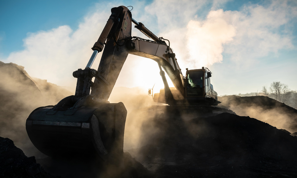An excavator among mounds of dirt, backlit by the sun and surrounded in plumes of dust from the dirt it is scooping.