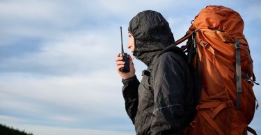 An individual in a black hooded jacket carrying a large orange backpack while using a black two-way radio outdoors.