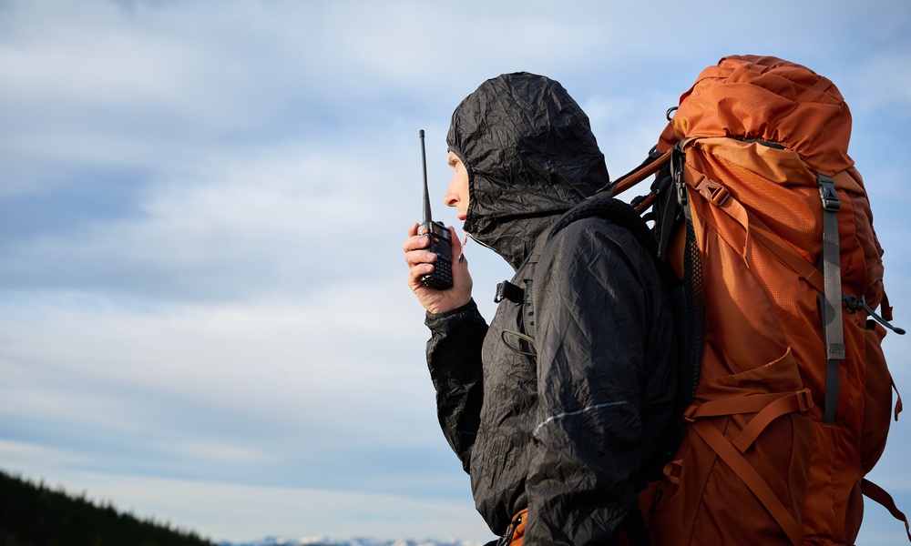 An individual in a black hooded jacket carrying a large orange backpack while using a black two-way radio outdoors.