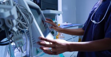 A nurse practitioner standing in a hospital operating room while using a monitor and other medical equipment.