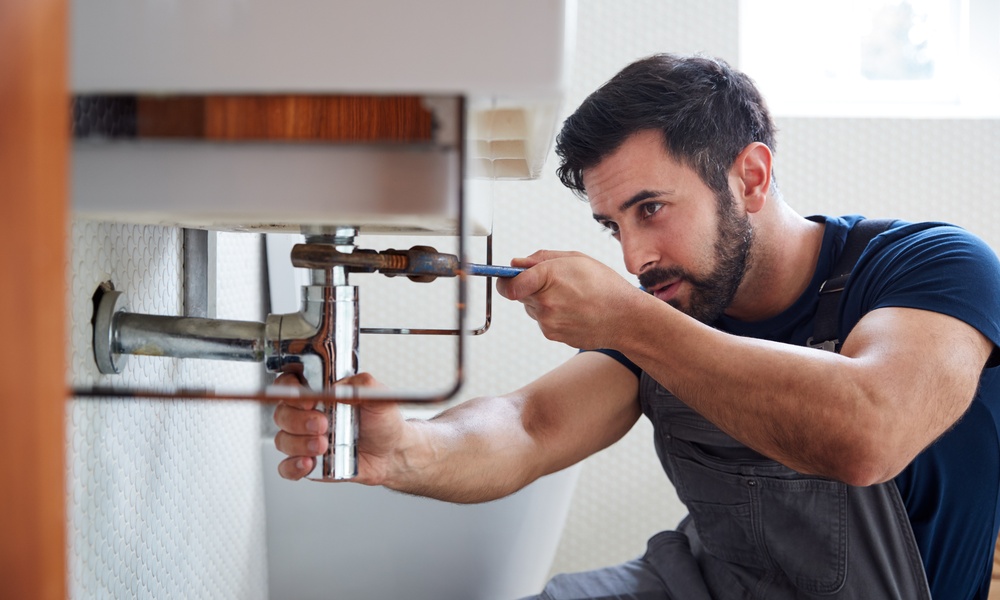 A close-up of a bearded man wearing a blue T-shirt and using a wrench to fix a pipe underneath a white sink.