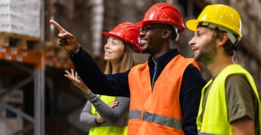 Three people wearing high-visibility vests and hard hats are standing in a warehouse as one person points a finger ahead.