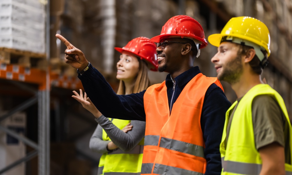Three people wearing high-visibility vests and hard hats are standing in a warehouse as one person points a finger ahead.
