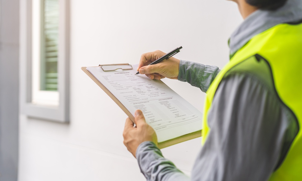 A person wearing a yellow safety vest fills out a form on a clipboard while examining the inside of a building.