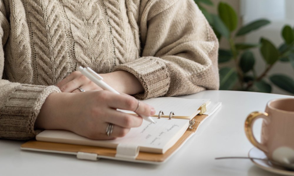 A woman wearing a sweater is sitting at a table with a mug. She's smiling as she holds a pen over a notebook.