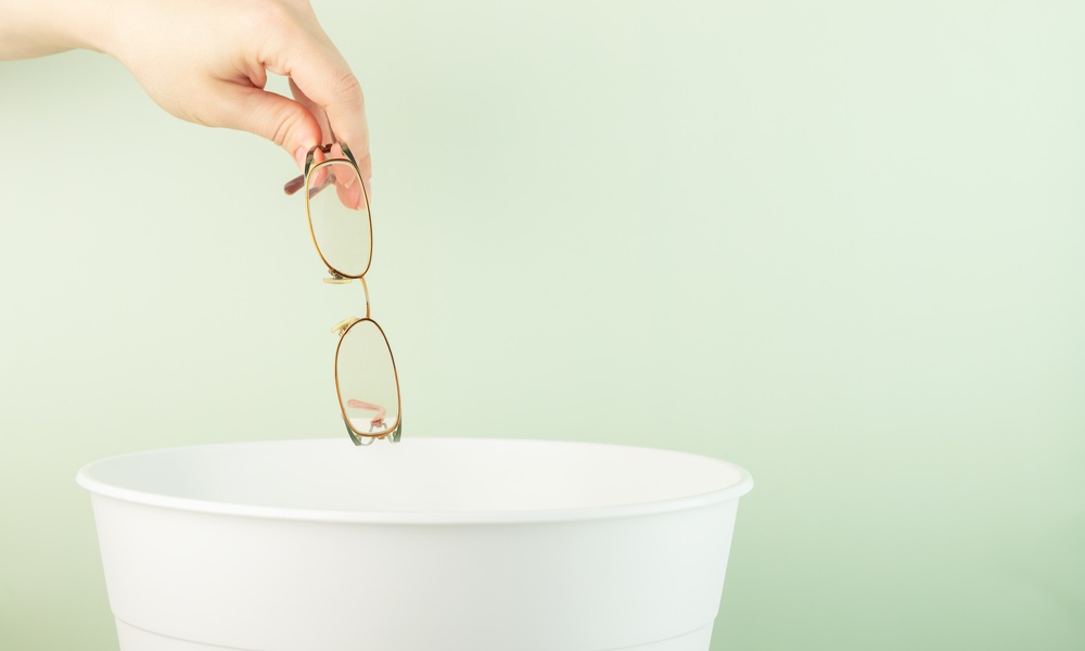 A person holding a pair of wire-frame glasses over a white trash bin, as if preparing to throw them away.