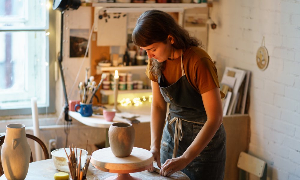 A person in a dark apron working with clay in a small studio space. There is an unfinished clay pot sitting on a wheel.