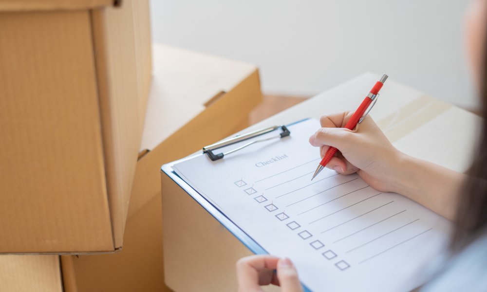 A woman surrounded by sealed cardboard boxes is holding a red pen and a clipboard with a checklist on it.