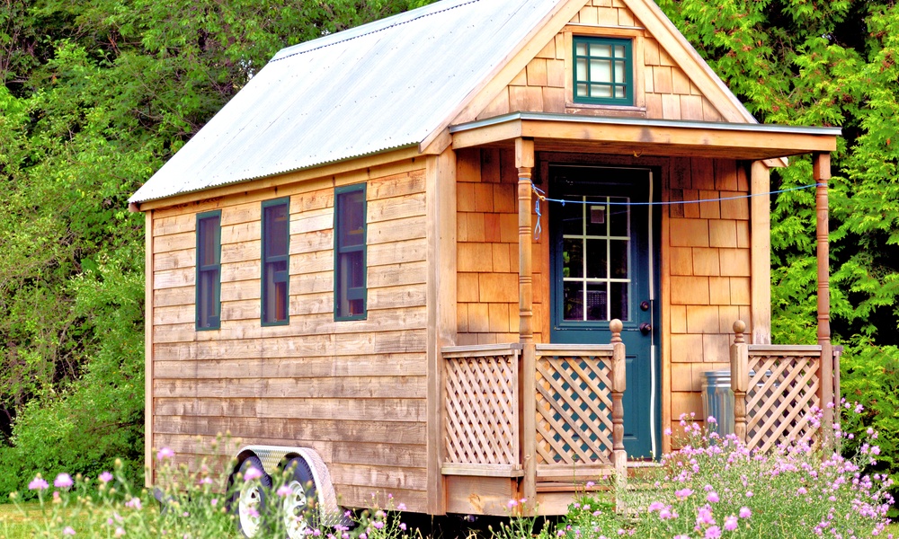 The exterior of a tiny home with wood siding, a metal roof, and a front porch. The home is on wheels and surrounded by trees.