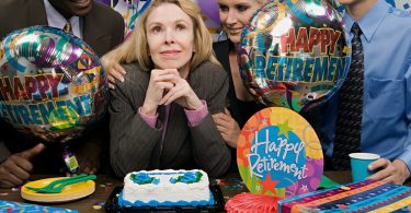 A woman dressed in professional attire sitting in front of a cake, balloons, and gifts at a retirement party with colleagues.