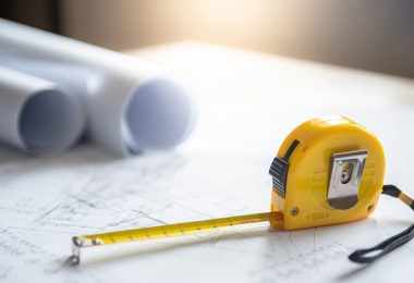 An extended yellow tape measure resting on top of a white engineering draft. Two rolled-up drafts sit nearby.
