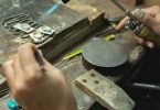 A person holding crafting tools in both of their hands against the surface of a workbench to create silver pieces.