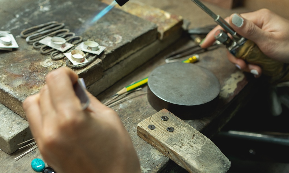 A person holding crafting tools in both of their hands against the surface of a workbench to create silver pieces.