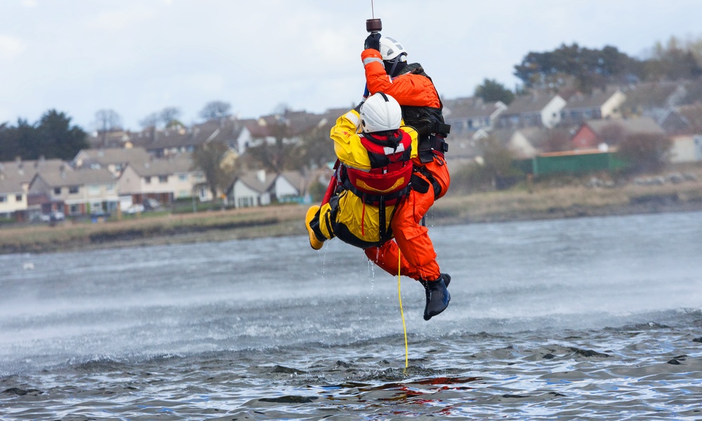 A water rescue mission being conducted with two individuals in white helmets hanging from a rope over the water.