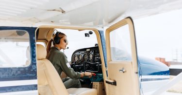 A woman pilot steers a white and blue private plane from the cockpit while wearing a headset and microphone.