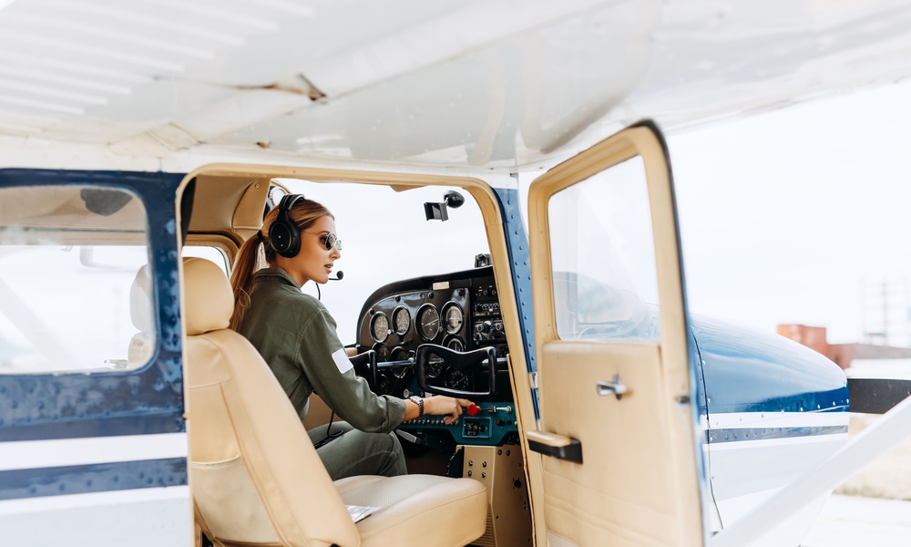 A woman pilot steers a white and blue private plane from the cockpit while wearing a headset and microphone.