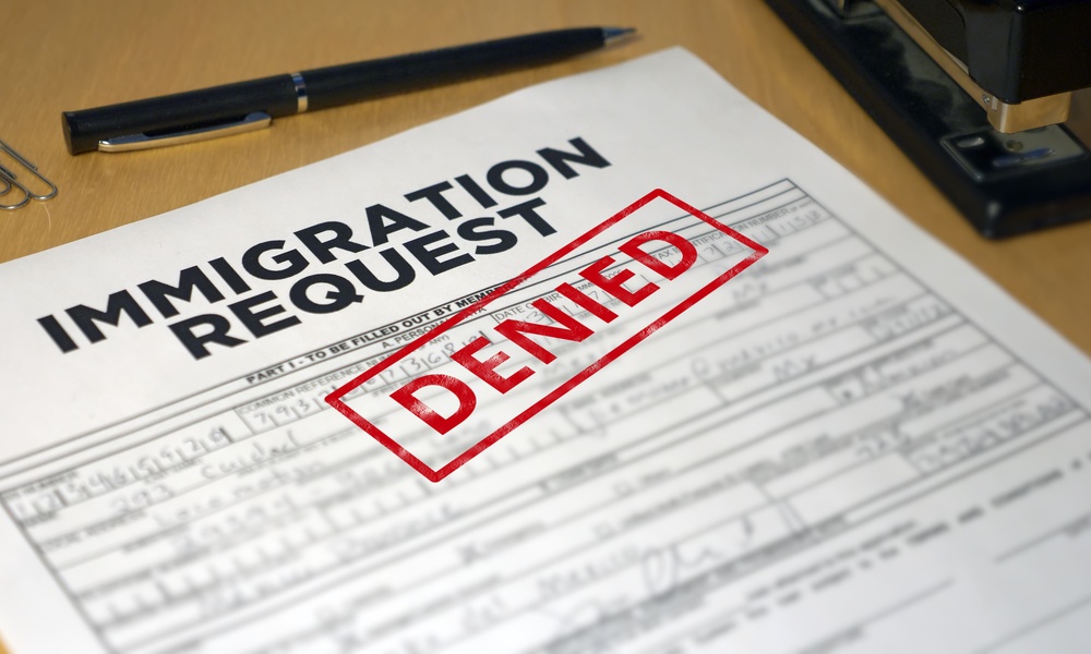 A document that says "Immigration Request" and "Denied" sitting on top of a wooden table next to a black pen.