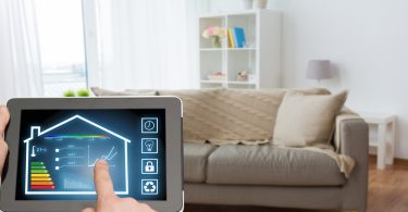 A person holding a tablet operating a smart home app while standing in their living room in front of a couch.