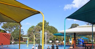 An outdoor circular pool with colorful umbrellas around the perimeter and a playground in the background.