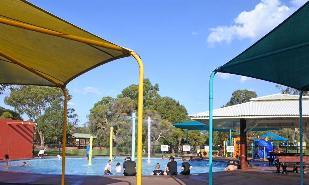 An outdoor circular pool with colorful umbrellas around the perimeter and a playground in the background.