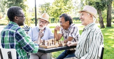 A group of four older men smiling and laughing while sitting at a table playing chess in a park on a sunny day.
