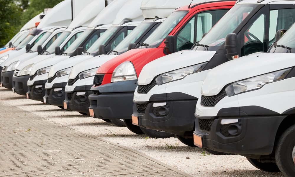 A row of parked commercial vans. They are all white, except for a single red van standing out among them on a paved lot.