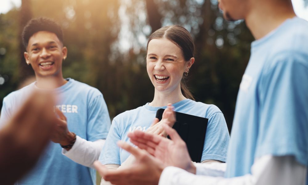 Three volunteers in pale blue T-shirts stand in front of trees, with the person in the middle holding a tablet.
