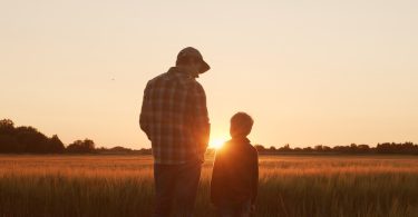 An adult farmer with a young boy standing in the middle of the field. They're watching the sun go down in front of them.