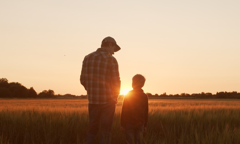 An adult farmer with a young boy standing in the middle of the field. They're watching the sun go down in front of them.